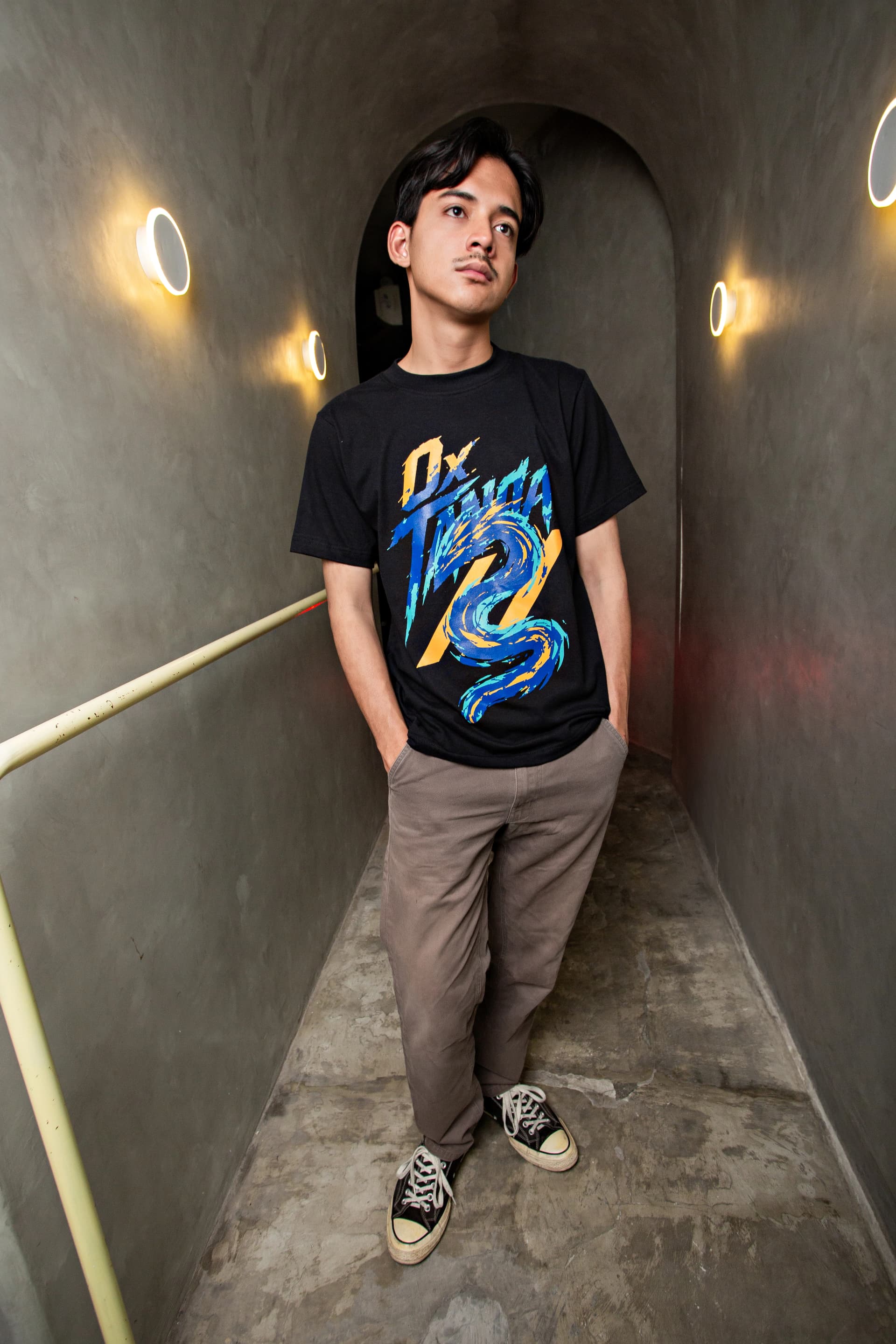 Man standing in a lit concrete corridor wearing a black graphic T-shirt
