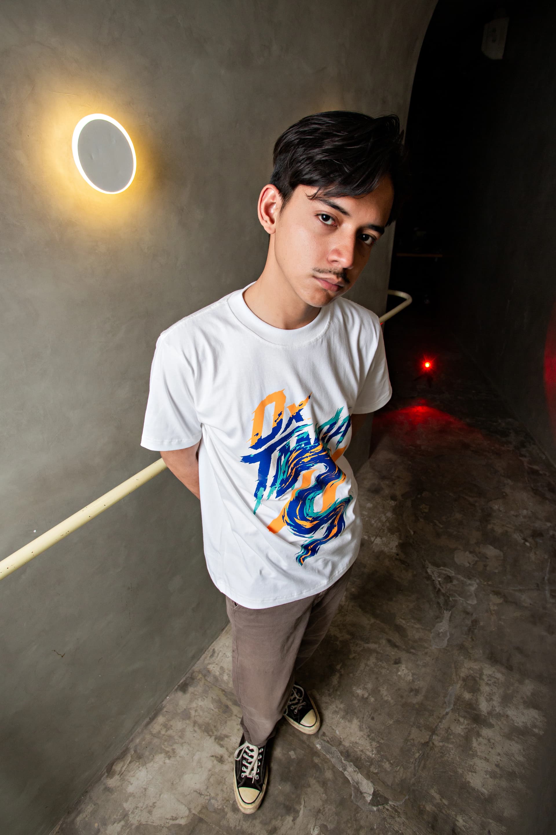 Man standing in a concrete corridor wearing a white graphic T-shirt and gray trousers